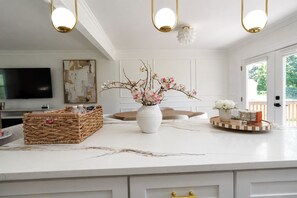 A modern kitchen with a marble countertop, a white vase with flowers, a woven basket, and a round wooden tray with items. - Stylish 4BR Boutique Home with Movie Lounge (Marietta)
