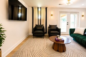 Een woonkamer met een flatscreen televisie, twee leren fauteuils, een groene bank, een ronde houten salontafel en een vloerkleed met dessin.