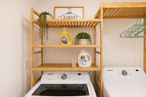 Een wasruimte met twee Samsung wasmachines, houten planken met wasmiddel, een potplant en een kledingrek.