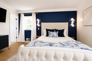 A bedroom with a tufted headboard, a bed with a floral-patterned bedspread, two bedside tables, and a wall-mounted light fixture. - Stylish 4BR Boutique Home with Movie Lounge (Marietta)
