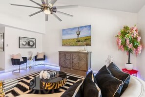 Un salon moderne avec un ventilateur de plafond, un tapis à motifs noir et blanc, une table basse ronde, un canapé avec des coussins noirs, un meuble, un tableau encadré et une plante en pot.