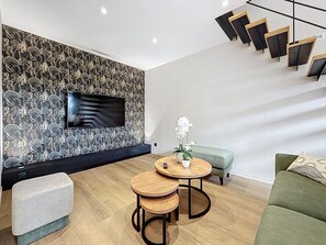 A modern living room with a flat-screen TV mounted on a decorative wall, two round wooden tables, a green sofa, and a staircase with wooden steps. - Vacation rental with a hot tub located between Saumur and Chinon, accommodating 14 people (Varennes-sur-Loire)