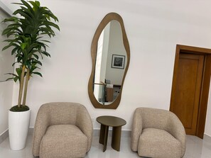 Ein Zimmer mit zwei Sesseln, einem kleinen Tisch, einem großen Spiegel und einer Zimmerpflanze.