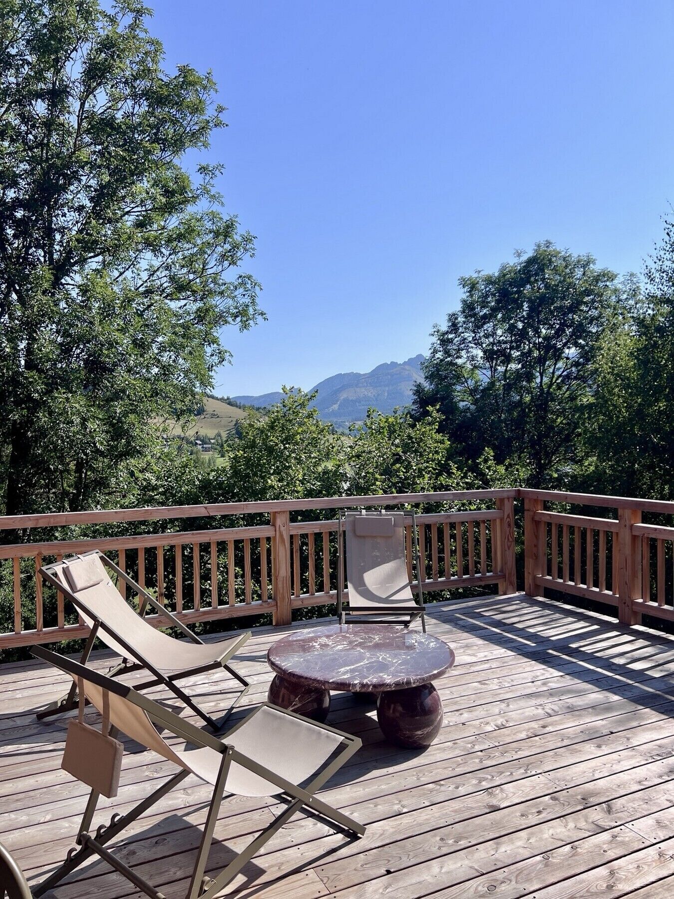 A wooden deck with two lounge chairs and a round table, overlooking a mountainous landscape.