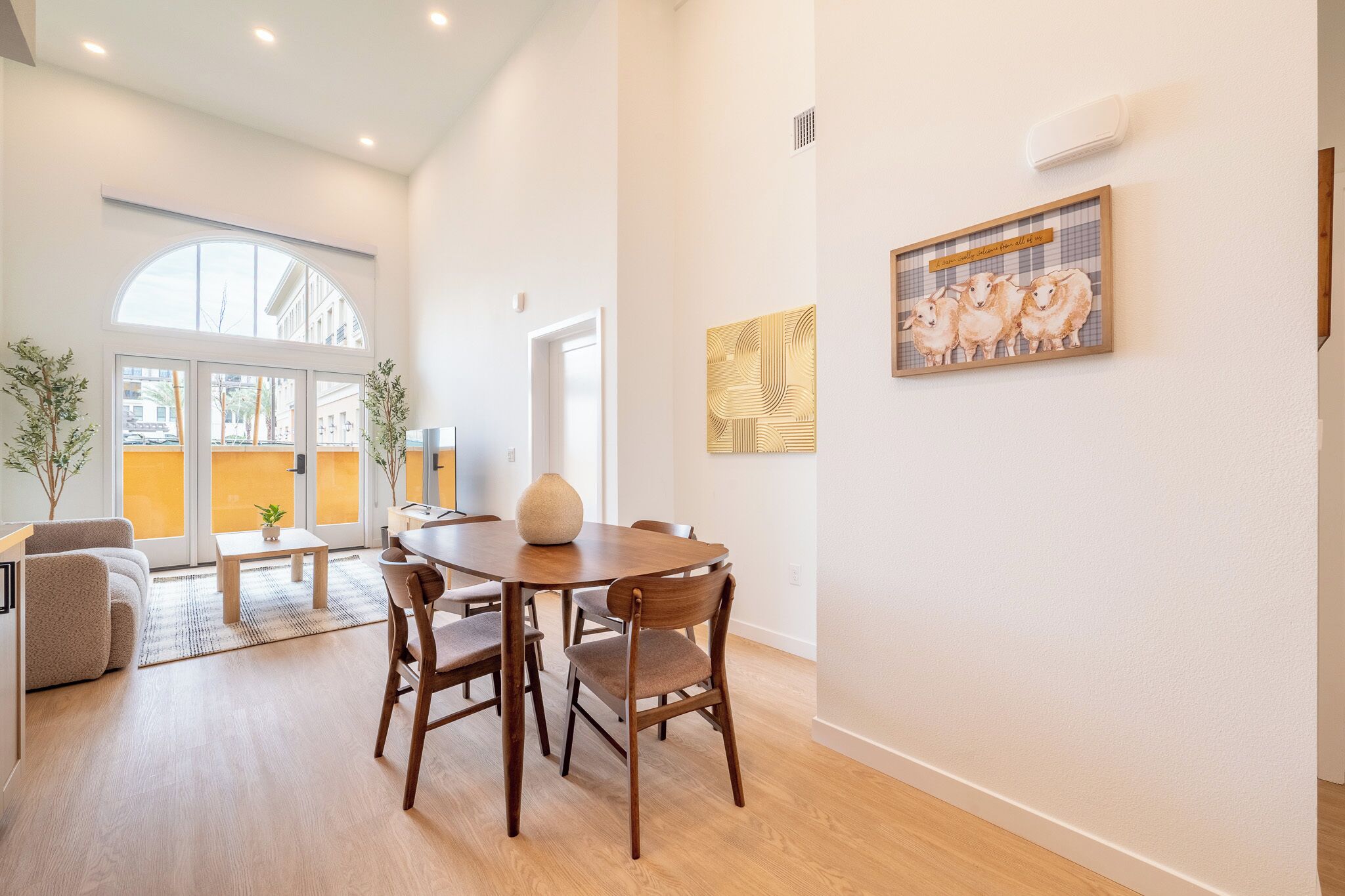 A spacious living area with a dining table, chairs, and a sofa. There is a large window providing natural light and a view of the outside.