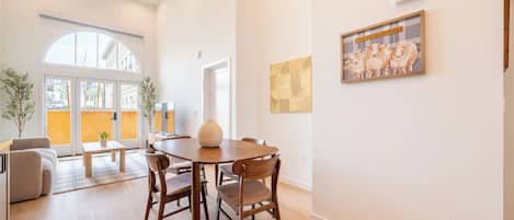 A spacious living area with a dining table, chairs, and a sofa. There is a large window providing natural light and a view of the outside.