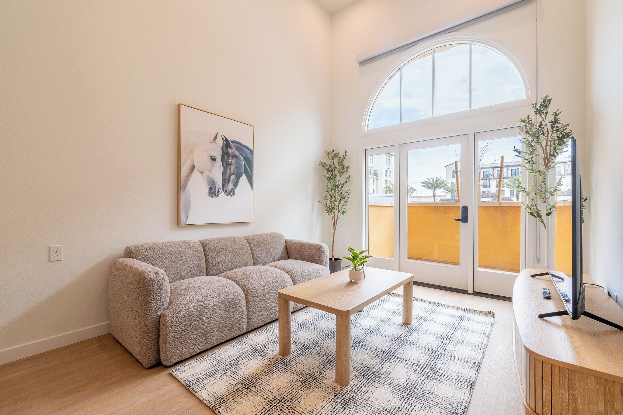 A living room with a beige sofa, a wooden coffee table, a framed horse painting, a potted plant, and a view through a glass door.
