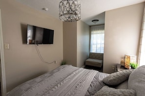 Quarto com cama, televisão na parede, uma cadeira e janela com cortinas.