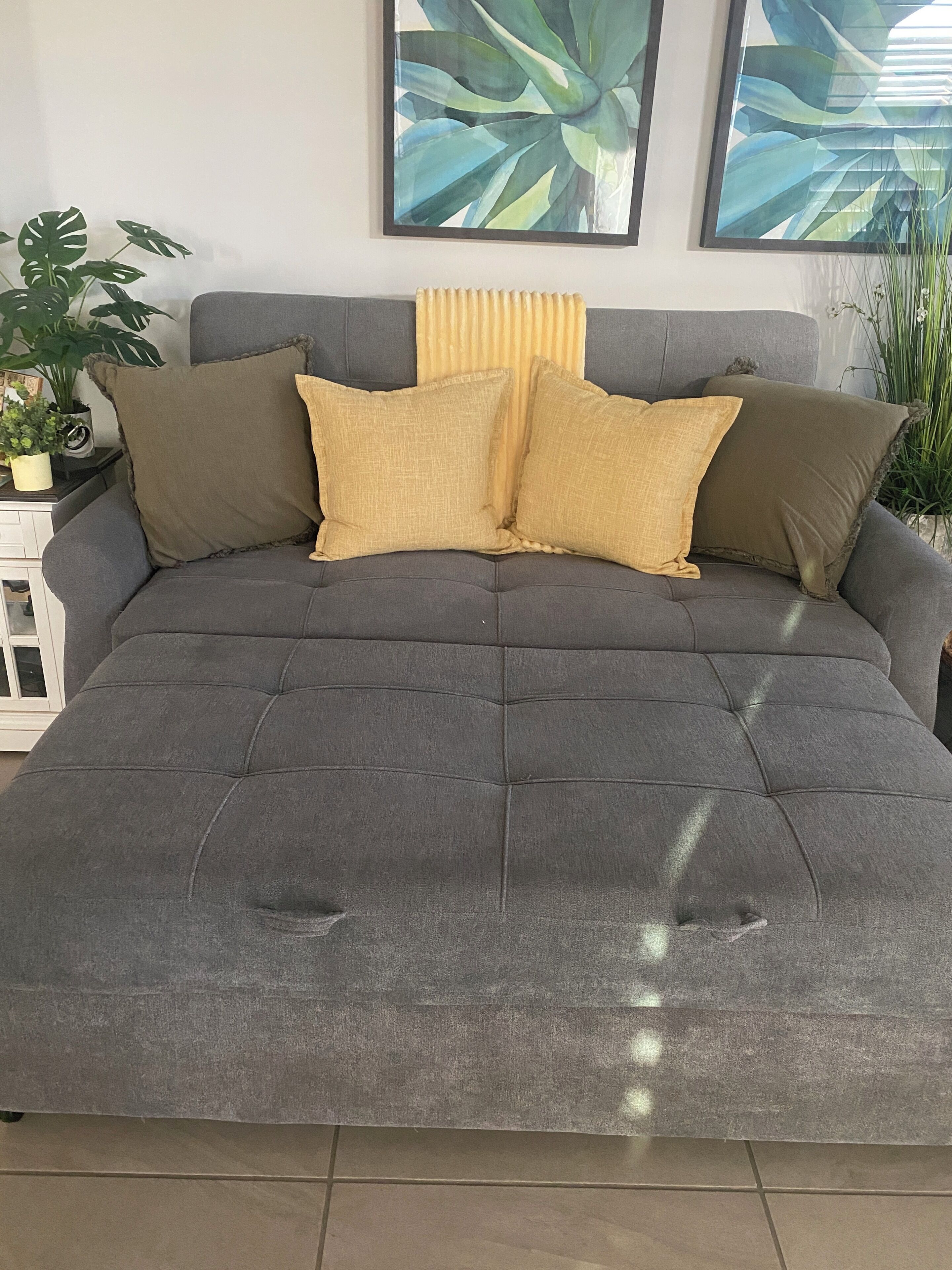 A grey upholstered sofa with a tufted design, accompanied by two matching cushions, set against a white wall with two framed botanical prints.