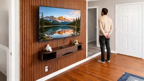 A person standing in a room with a wooden wall paneling, a mounted television displaying a scenic landscape, and a white door in the background. - Urban Oasis | Stylish 2BR Near Med District w/ Pool & Free Parking (Dallas)