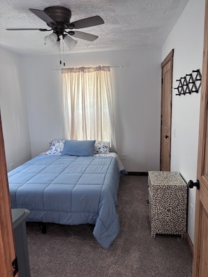 A bedroom with a bed, a ceiling fan, a window with curtains, a door, and a small cabinet.