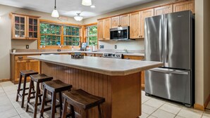 A kitchen with wooden cabinets, a stainless steel refrigerator, a microwave, and a large island with stools. - Lakefront on Winnipesaukee - Private Dock, Beach & Firepit - Sleeps 10 (Laconia)