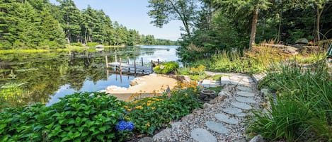 A serene lakeside pathway with a dock, surrounded by lush greenery and a variety of plants.