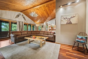 A spacious living room with a high wooden ceiling, a large sectional sofa, a coffee table, and a painting of a deer on the wall. - Lakefront on Winnipesaukee - Private Dock, Beach & Firepit - Sleeps 10 (Laconia)