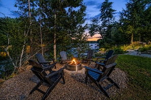 An outdoor seating area with Adirondack chairs around a fire pit, overlooking a body of water and a forested area. - Lakefront on Winnipesaukee - Private Dock, Beach & Firepit - Sleeps 10 (Laconia)