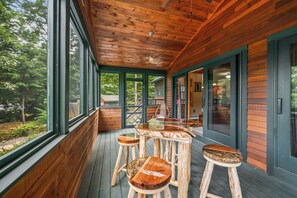 A wooden deck with a table and stools, large windows, and a view of the surrounding forest. - Lakefront on Winnipesaukee - Private Dock, Beach & Firepit - Sleeps 10 (Laconia)