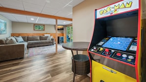 A room with a Pac-Man arcade machine, a sofa, a wicker table, and a window with a view of greenery. - Lakefront on Winnipesaukee - Private Dock, Beach & Firepit - Sleeps 10 (Laconia)