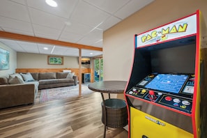Ein Zimmer mit einem Pac-Man-Spielautomaten, einer Couch, einem Korbtisch und einem Fenster mit Blick auf Grünflächen.