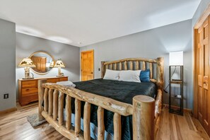 Een slaapkamer met een houten hoofdeinde, een bed met kussens, een dressoir met lampen, een spiegel en een deur.