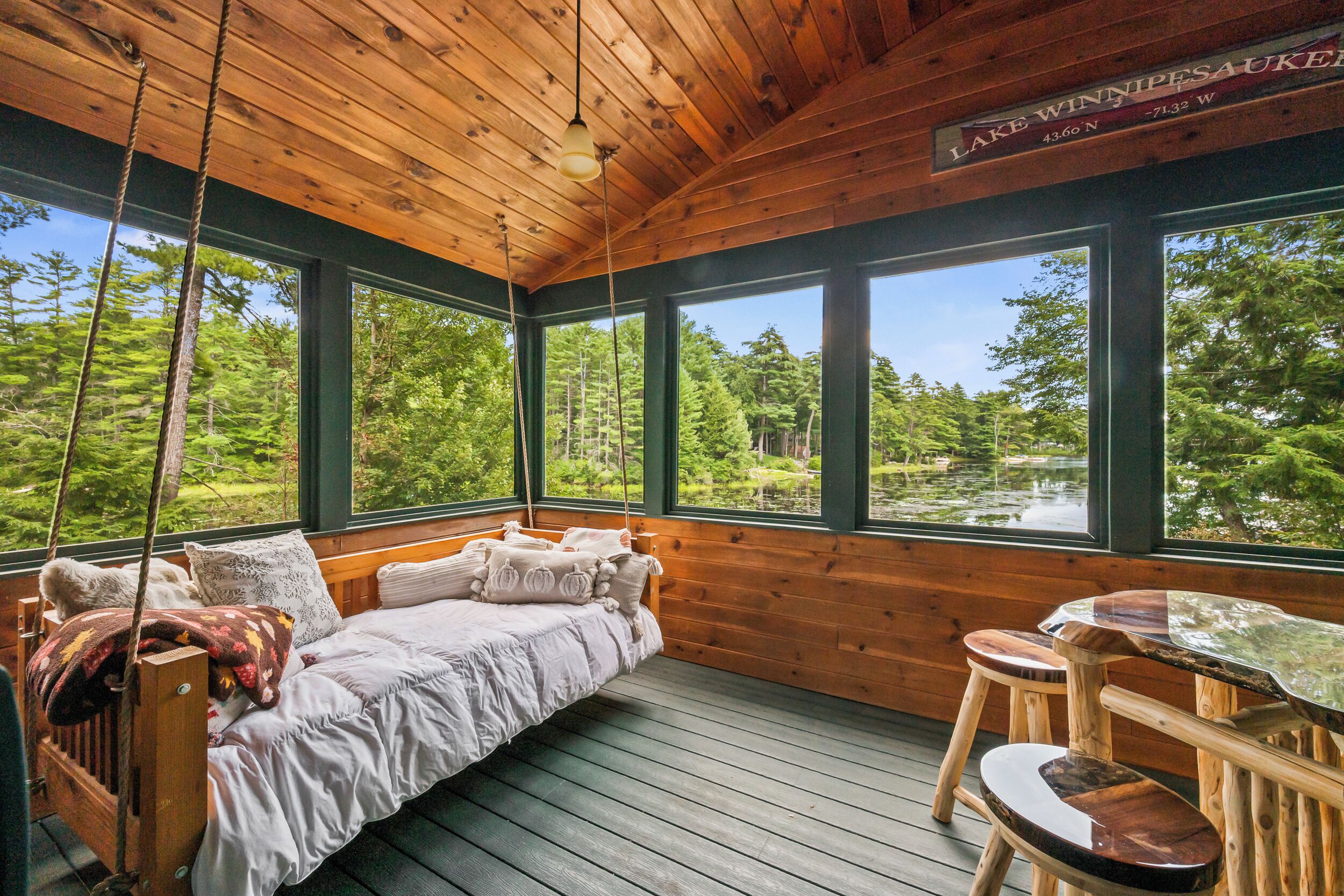 A cabin with a wooden interior, a bed with pillows, a table, and large windows overlooking a forest and a lake.