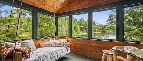 A cabin with a wooden interior, a bed with pillows, a table, and large windows overlooking a forest and a lake.