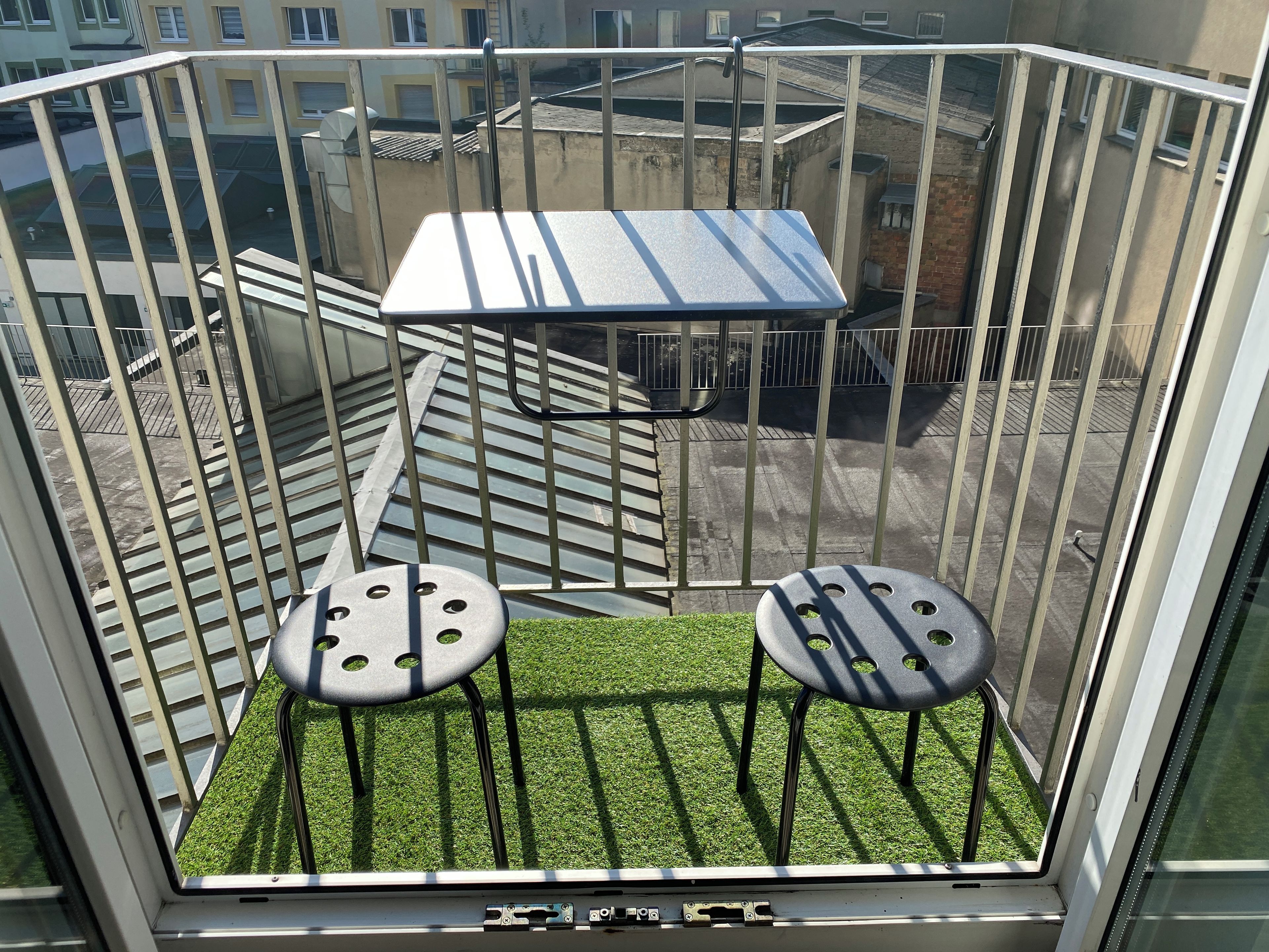 Ein Balkon mit Metallgeländer, zwei Hocker, einem kleinen Tisch und Blick auf Gebäude.
