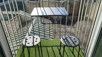 Ein Balkon mit Metallgeländer, zwei Hocker, einem kleinen Tisch und Blick auf Gebäude.