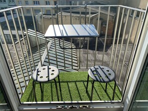 Ein Balkon mit Metallgeländer, zwei Hocker, einem kleinen Tisch und Blick auf Gebäude.