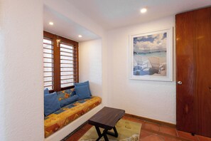 Ein Zimmer mit Holztür, einem Fenster mit Jalousien, einer Couch mit Kissen, einem gerahmten Bild an der Wand und einem Fliesenboden.