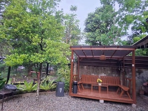 A covered outdoor seating area with a bench and table, surrounded by greenery and a concrete wall.
