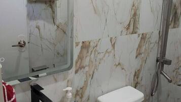 Un baño con pared de mármol, lavamanos blanco, inodoro y ducha. Hay una toalla colgada en la pared y un cartel con instrucciones.