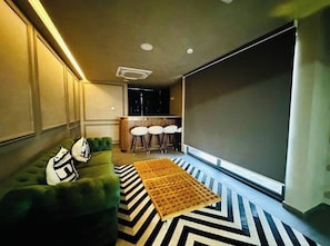 Un salon moderne avec un canapé vert, un tapis à motifs noir et blanc, un écran de projection et un coin bar avec des tabourets.