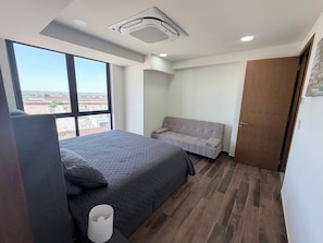 A bedroom with a bed, a grey sofa, a wooden door, and a window with a view.