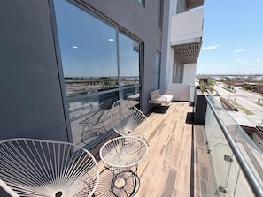 Un balcon offrant une vue sur le paysage urbain, avec un design moderne, une balustrade en verre, des fauteuils en osier et une petite table.