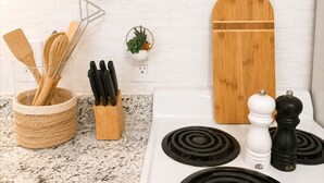 A kitchen with a white stove, black stovetop burners, a wooden cutting board, a spice rack with white and black pepper shakers, and a woven basket holding wooden utensils.