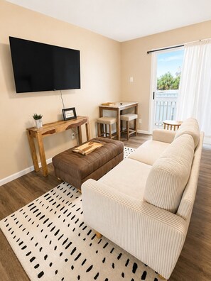 Una sala de estar con un sofá, una mesa de centro, un televisor montado en la pared, una pequeña mesa de madera con una planta, y vistas al exterior a través de una puerta corredera.