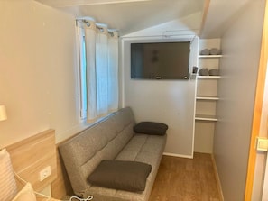 Ein kleiner Raum mit einer grauen Couch, einem an der Wand montierten Fernseher, einem Fenster mit Vorhängen und einem Holzboden.