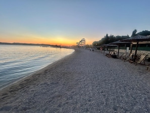 Ein Kiesstrand mit einer Reihe von Liegestühlen und einer überdachten Sitzgelegenheit.