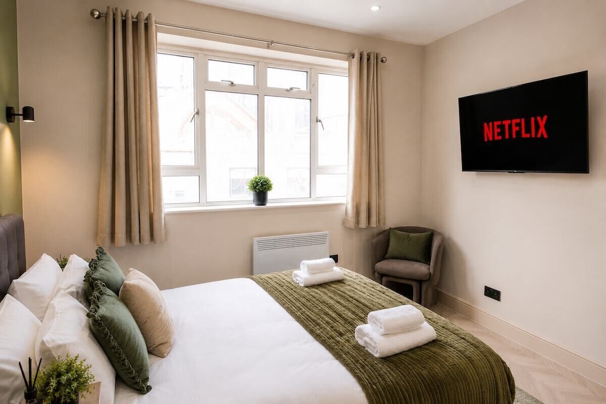 Habitación de hotel con cama, televisor montado en la pared, ventana con cortinas, radiador y una pequeña planta en el alféizar.