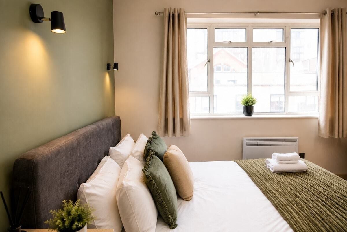 Habitación de hotel con cama, almohadas, mesita de noche con una planta, ventana con cortinas y lámpara de pared.