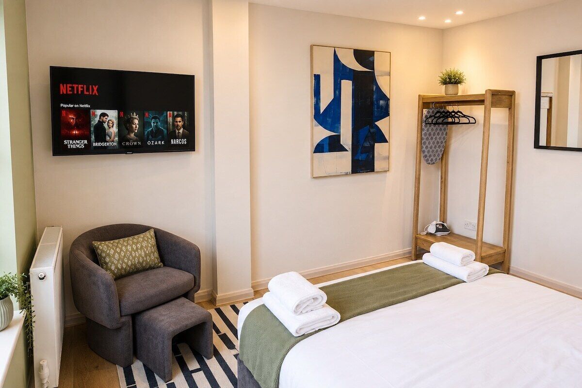 A hotel room with a bed, a chair, a wall-mounted TV displaying Netflix shows, a painting, and a mirror.