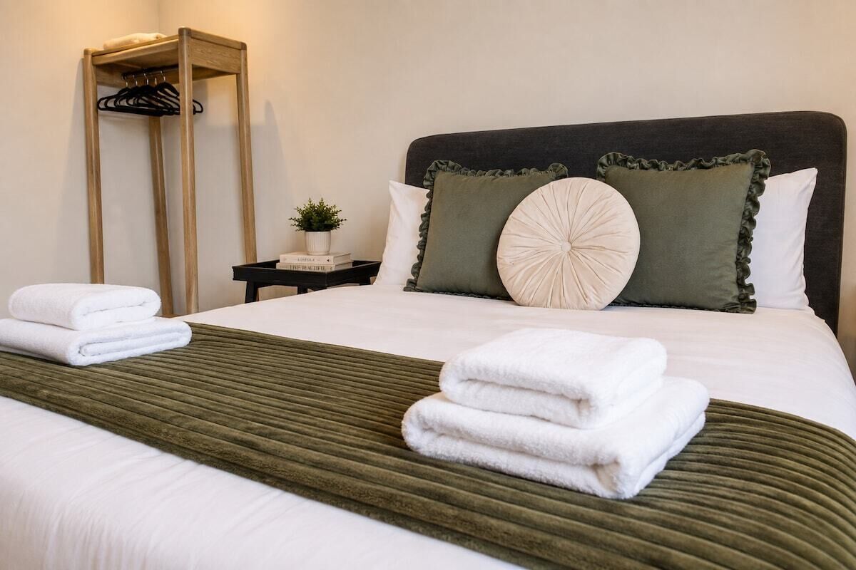 Ein ordentlich bezogenes Bett mit weißen Laken und einer grünen Tagesdecke, ein Nachttisch mit Pflanze sowie ein hölzerner Kleiderständer.