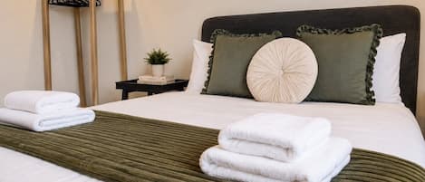 A neatly made bed with white linens and a green throw blanket, a bedside table with a plant, and a wooden coat rack.