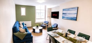 En moderne stue med en sofa, et spisebord dækket til fire personer, og et fjernsyn ophængt på væggen.