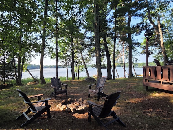 A lakeside seating area with Adirondack chairs and a wooden bench, surrounded by trees and a view of the lake.