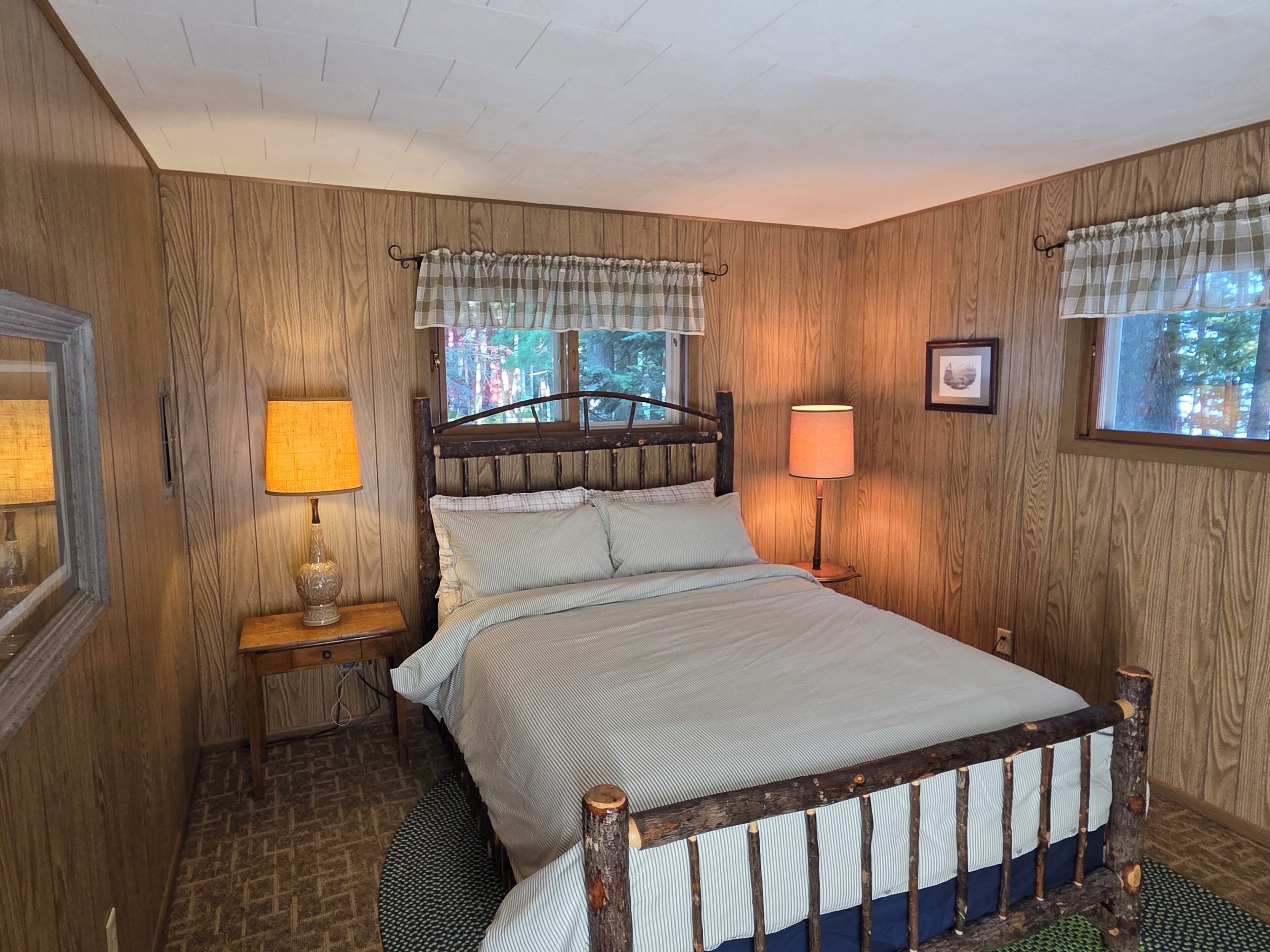 Une chambre à coucher avec une tête de lit en bois, un lit avec une literie à rayures, deux lampes de chevet et une fenêtre donnant sur des arbres.