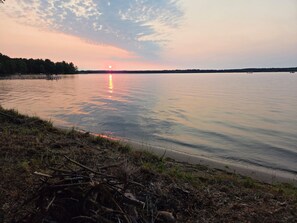 Un lago tranquilo al atardecer con cielo despejado, un muelle a la izquierda y una orilla con vegetación.