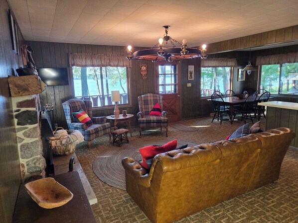 Una sala de estar acogedora con un sofá de cuero, sillones de cuadros, una chimenea de piedra y una zona de comedor con mesa y sillas de madera.