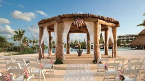 An outdoor wedding ceremony setup with a wooden arch, white chairs, and a white aisle.