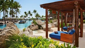 A resort pool area with a wooden gazebo, lounge chairs, and palm trees.
